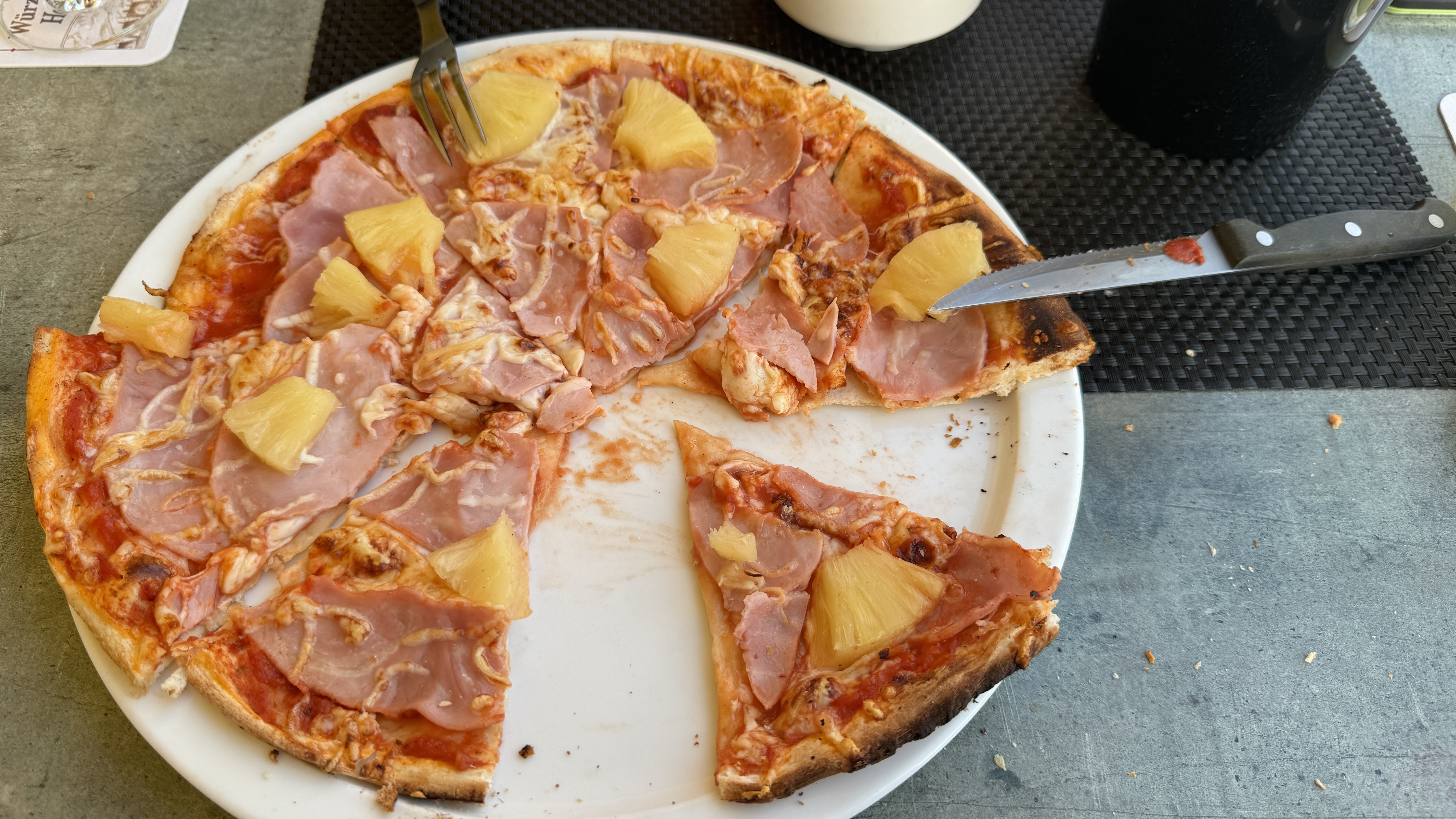 Bild: Auf einem Teller, Liegt eine Dreiviertel  Pizza und ein weiteres Stück dieser Pizza. Die Pizza ist mit Annanas und Schinken belegt.